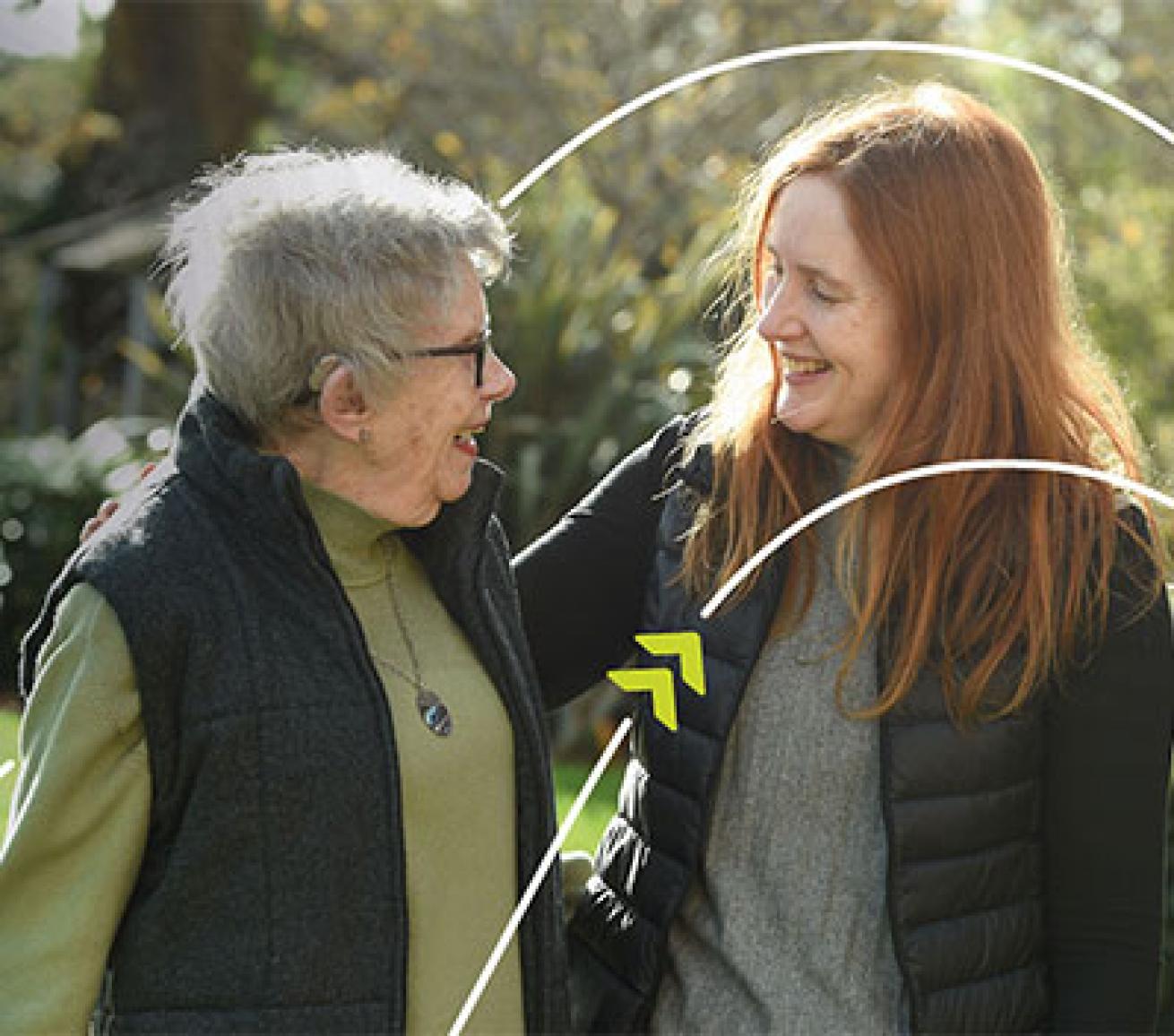 Two people standing together outdoors in a garden, with one person placing an arm around the other's shoulder.