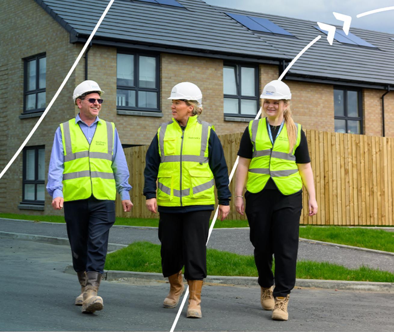 Three people wearing high-visibility clothing and hard hats walking through a residential housing development with solar panels on the roofs.