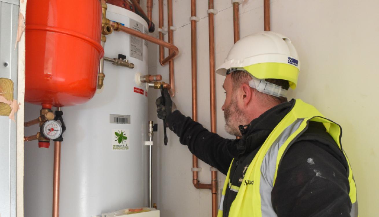 A technician in a safety helmet and high-visibility vest is inspecting a plumbing system.