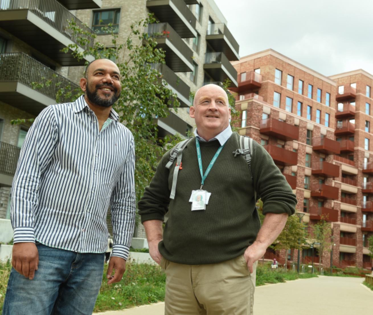 Two men stand outdoors in front of modern apartment buildings. The man on the left, wearing a striped shirt, smiles and looks towards the camera.