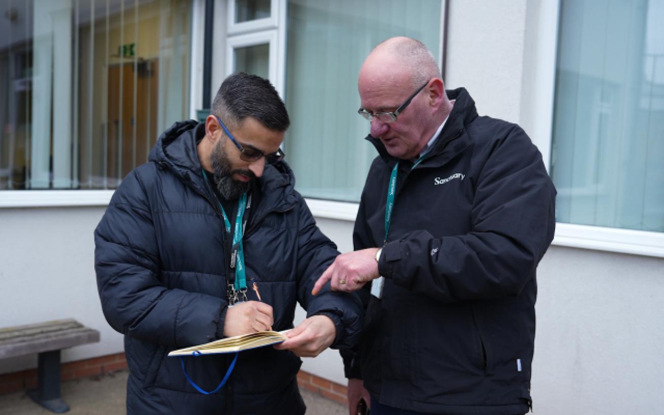 Two men are standing outside a building. The man on the left, wearing glasses and a dark puffer jacket, is writing on a notepad.