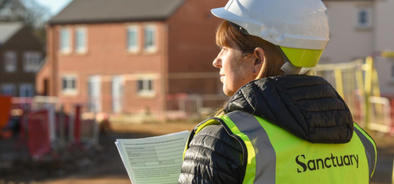 A woman wearing a white hard hat and a bright yellow safety vest with the word "Sanctuary" printed on the back stands on a construction site.