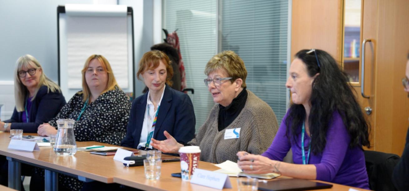A group of five women are seated around a conference table in a meeting room. They are engaged in discussion, with one woman speaking animatedly.