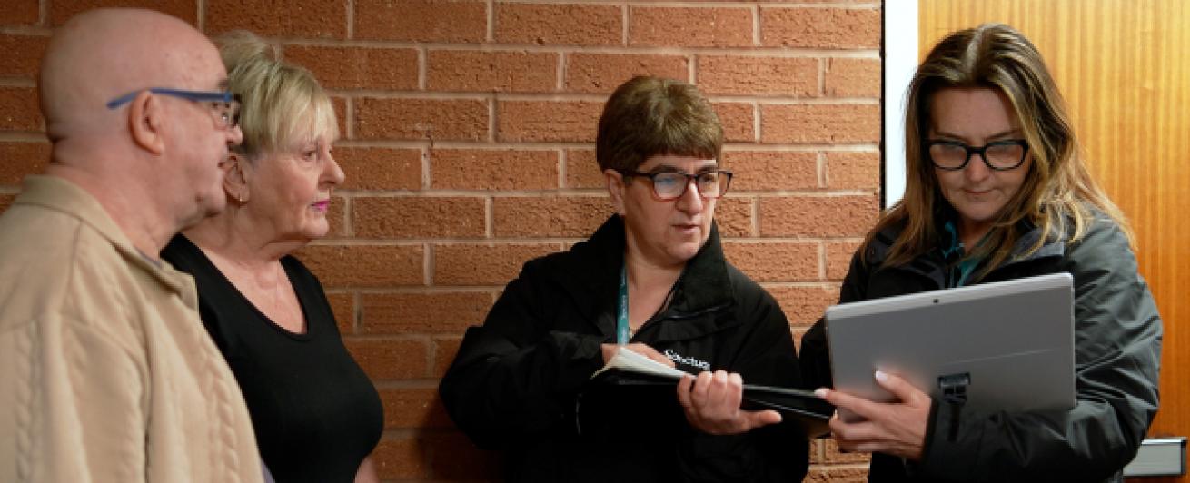A group of four people stand indoors near a brick wall. One person is holding a tablet and showing it to the others.