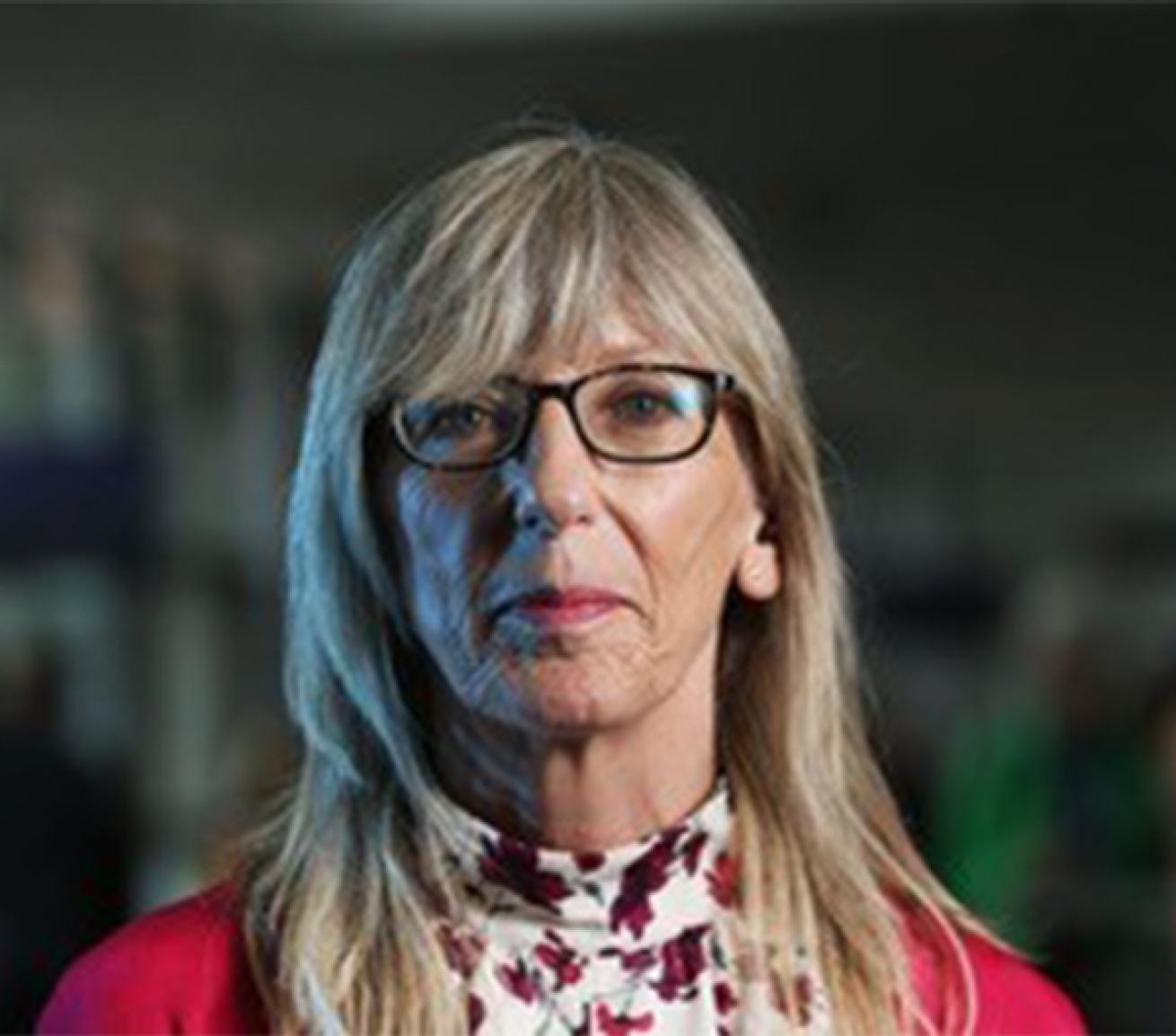 Head-and-shoulders portrait of a person with long grey hair and dark-framed glasses, wearing a white floral-patterned high-neck top and a red cardigan