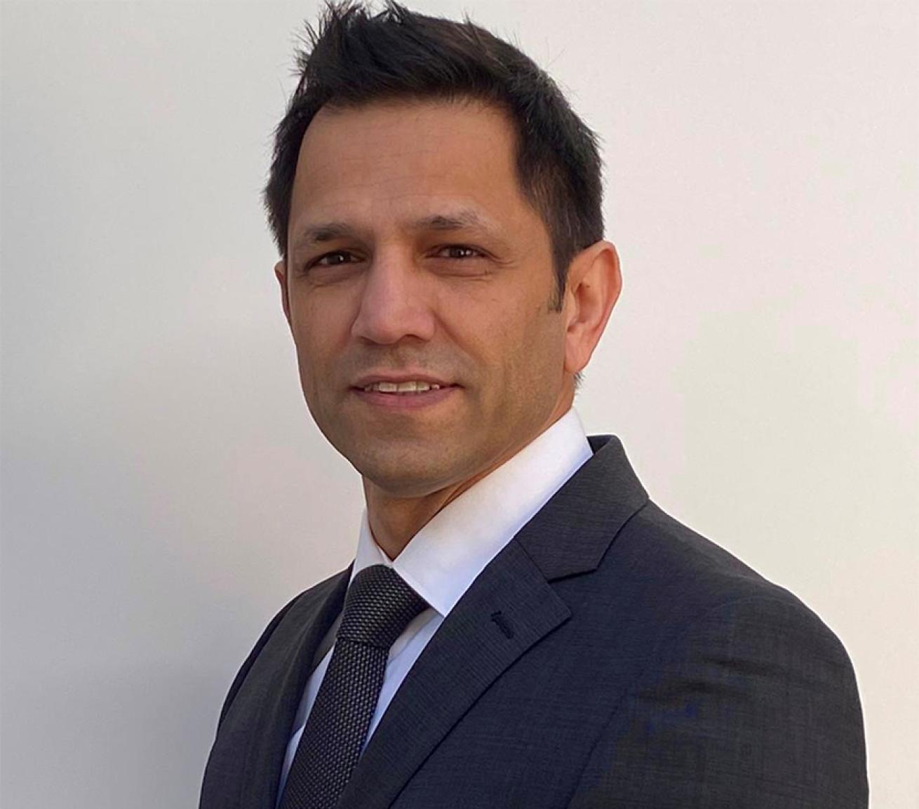 Portrait photograph of a person wearing a dark suit, white shirt and tie, positioned against a plain light background and looking towards the camera.