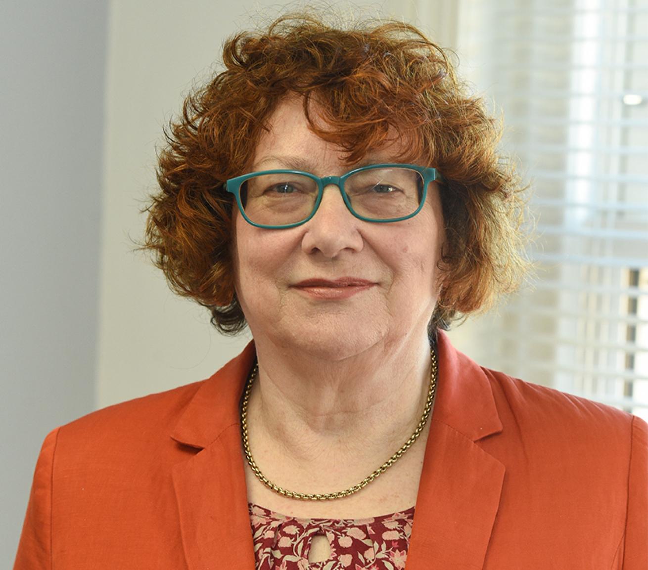 Portrait photograph of a person with short curly hair and glasses, wearing an orange blazer and patterned top, standing indoors with window blinds visible in the background.