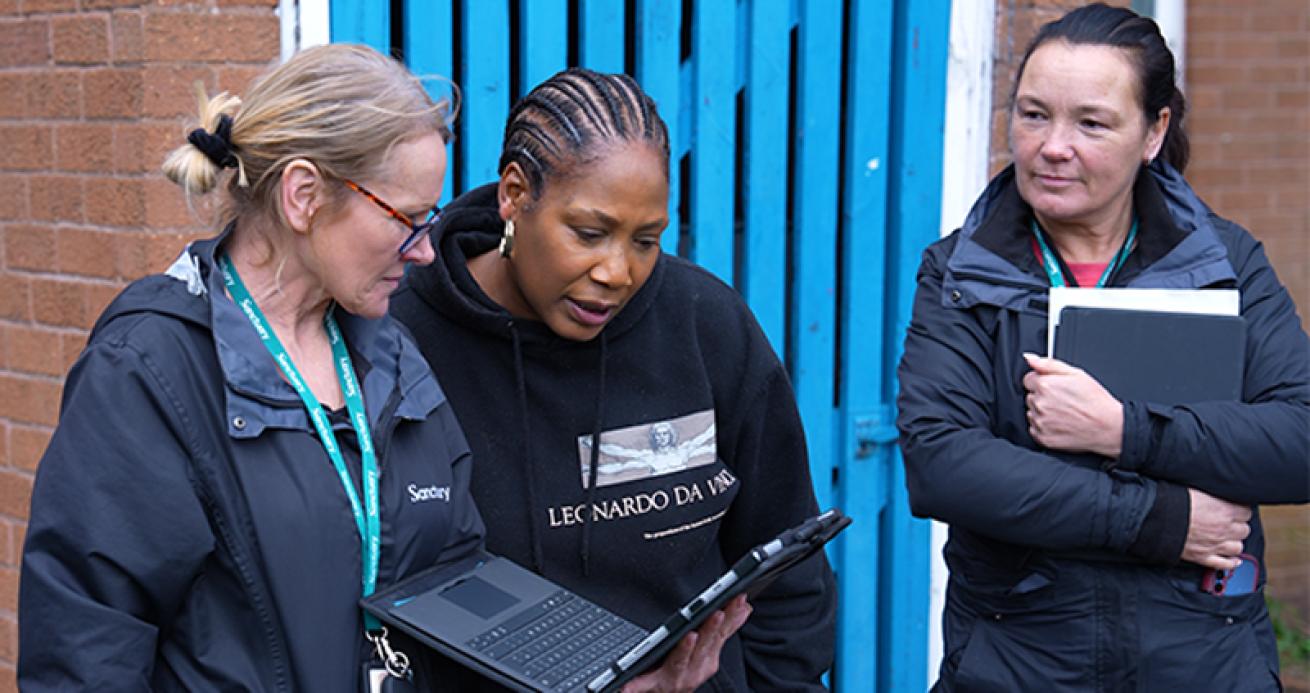 Three staff members standing outside a brick building review information on a tablet device. Two people look at the tablet while a third stands nearby holding a notebook.