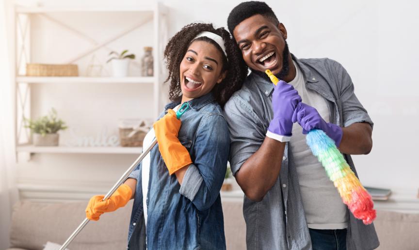A young woman and man leaning on each other smiling and holding a duster and a brush
