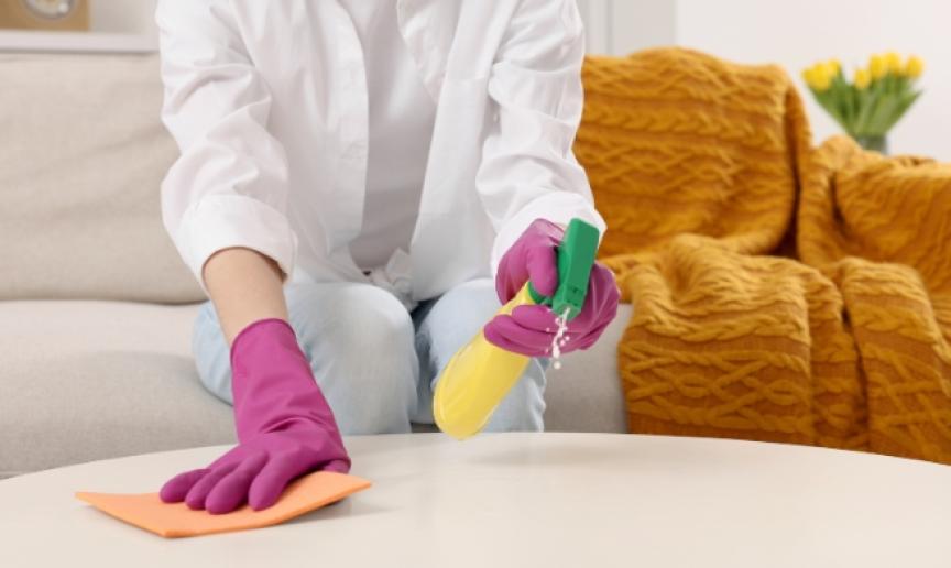 A person with yellow marigold gloves on holding cleaning spray and a sponge cleaning a wooden coffee table