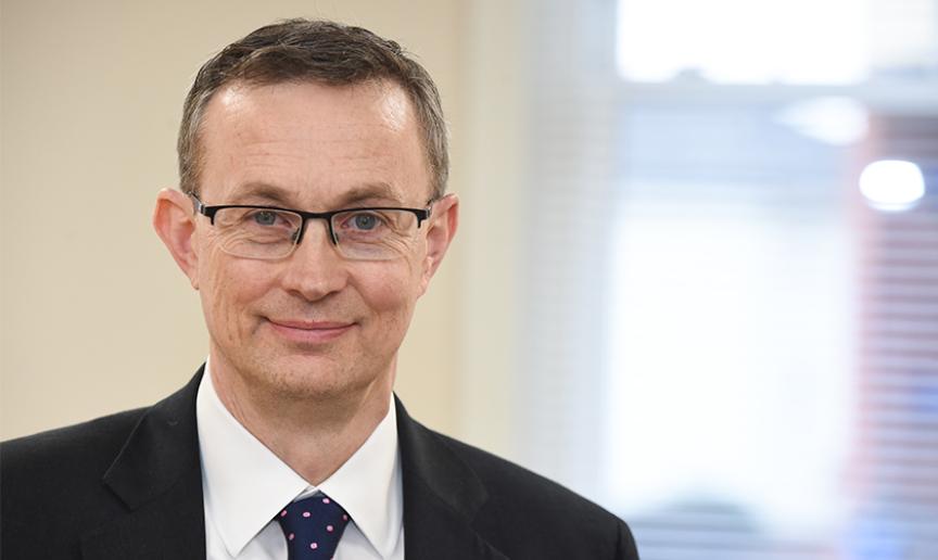 A man with brown hair wearing glasses, a black jacket, white tie and pink spotted tie looking at the camera and smiling