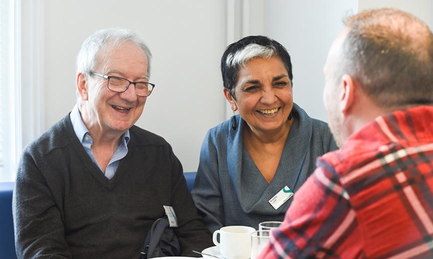 A woman with black and white hair wearing a grey top sitting on a blue bench next to a man wearing a blue shirt and grey jumper. Both people are wearing Sanctuary name badges and smiling at a man wearing a red checked shirt.