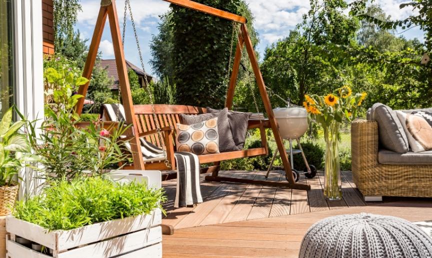 A wooden decking area with a swing seat, sunflowers, a barbecue and more plants 