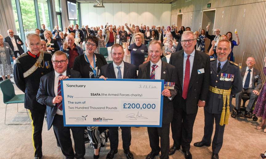 A large group of people standing in a room raising their glasses in celebration. In the forefront of the picture are six males and one female holding a large cheque for £200,000 from Sanctuary to the Armed Forces charity. Some of the men are wearing military medals.