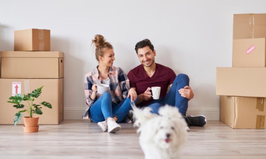 A couple sat on the floor of their new home with boxes beside them. They have a mug in their hand and look happy. Their small white dog is running towards the camera in front of them