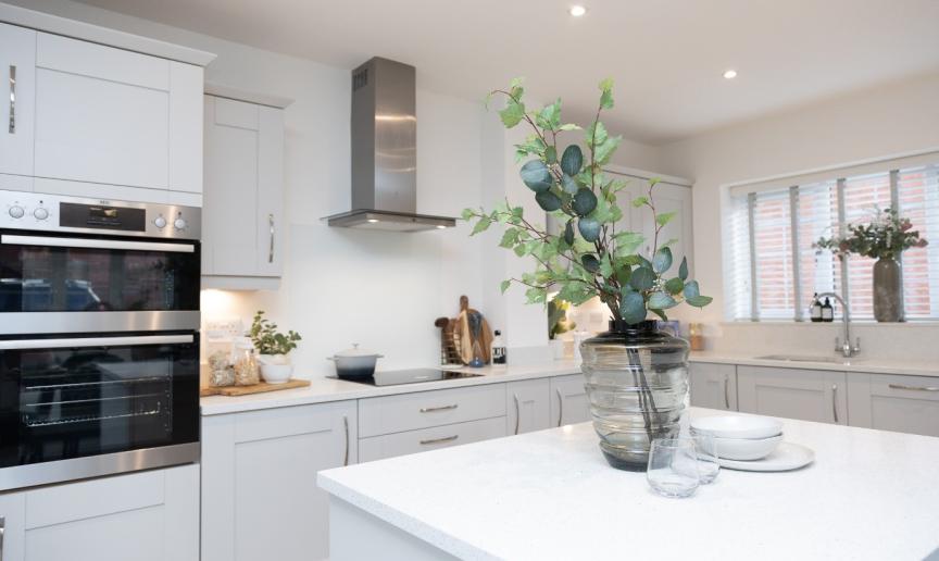 Interior of a clean and tidy white kitchen with cream cabinets. In the foreground is a kitchen island on which is a vase with foliage in and some crockery next to it