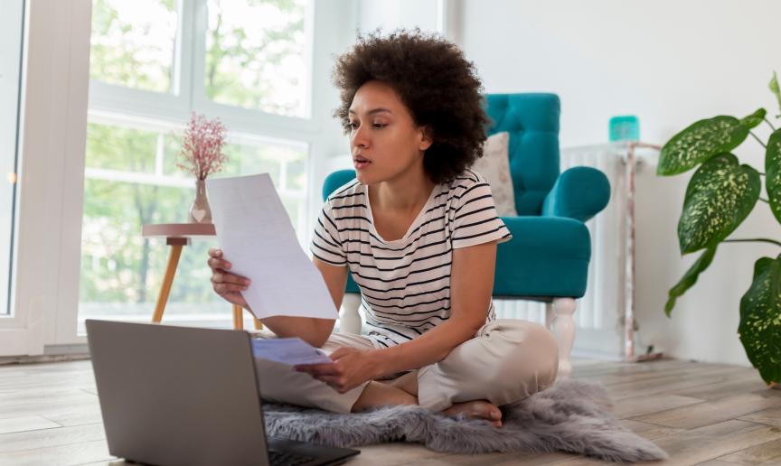 Stock image of a young woman sitting on a rug on a floor holding several pieces of paper and looking at a laptop