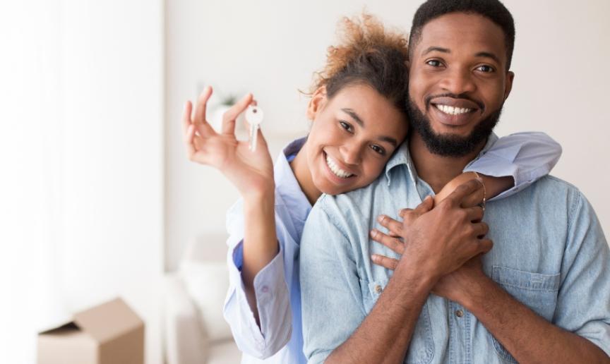 A male and female couple smiling at the camera, with the female holding up a key. She has her arm over the male in a sweet embrace