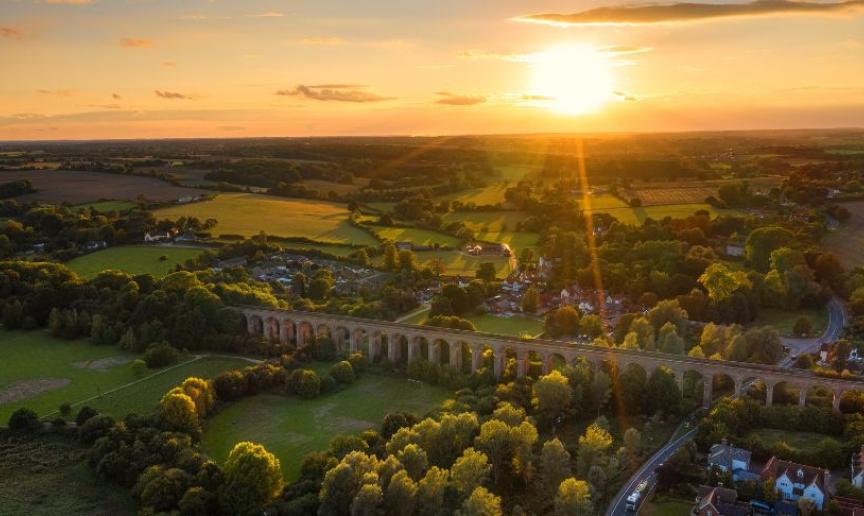 Aerial view of a countryside viaduct at sunset, with green fields, trees, and scattered houses.