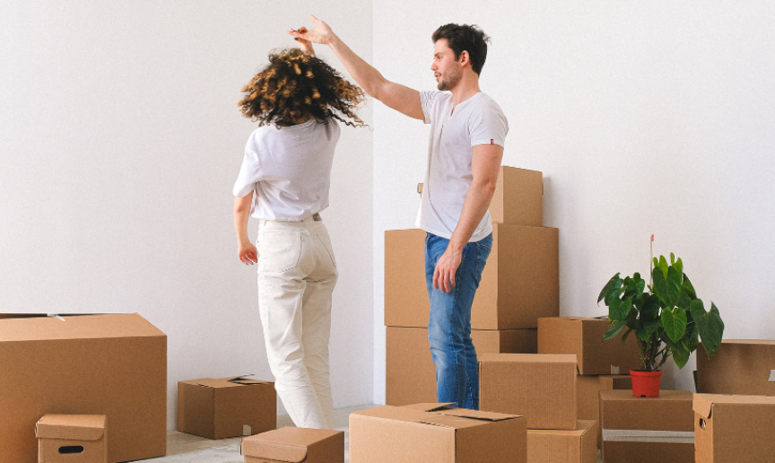 Two people in a room full of cardboard boxes, with one person playfully spinning the other by the hand. A green plant in a red pot is visible among the boxes.