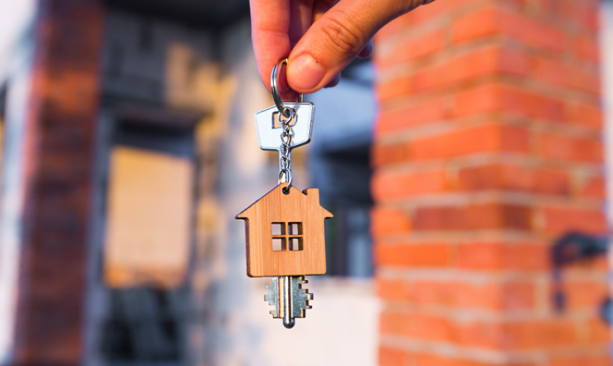 A house key with a home shaped wooden key ring attached are being held by two fingers. In the blurred background you can see a red bricked home.