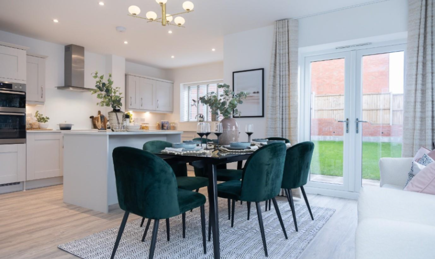 Open-plan kitchen/diner with white cabinets and quartz worktops. The dining space has a large table and 6 dark green velvet chairs, with a white sofa in the family area. Double doors open onto a rear garden.