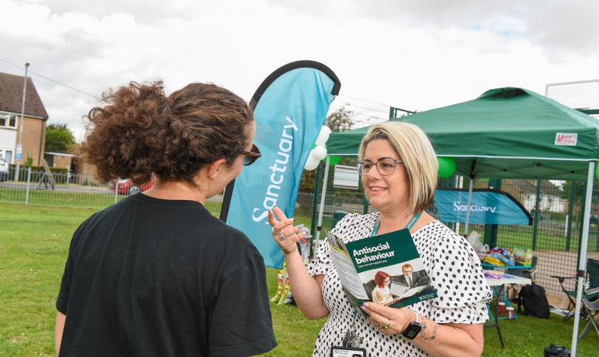 A woman with short blonde hair and glasses is speaking to a person with curly hair, wearing a black shirt and sunglasses. The woman is holding brochures titled "Antisocial behaviour" and is gesturing as she talks. In the background, there is a green tent with the word "Sanctuary" displayed on a banner, and various items are set up on a table. The setting appears to be an outdoor community event, with a grassy area and a fence in the background.