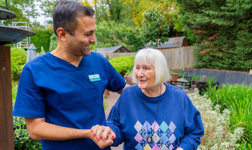 A healthcare worker in a blue uniform smiles and holds hands with an elderly woman in a blue sweater adorned with a diamond pattern.