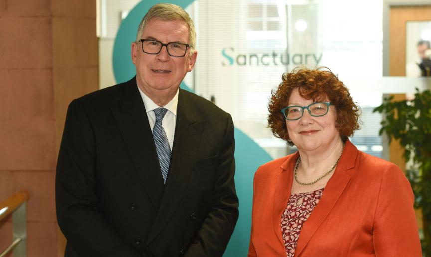 Two people standing side by side in an office reception area, with a large Sanctuary logo visible behind them.