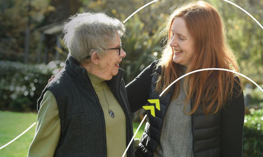 Two people standing together outdoors in a garden, with one person placing an arm around the other's shoulder.