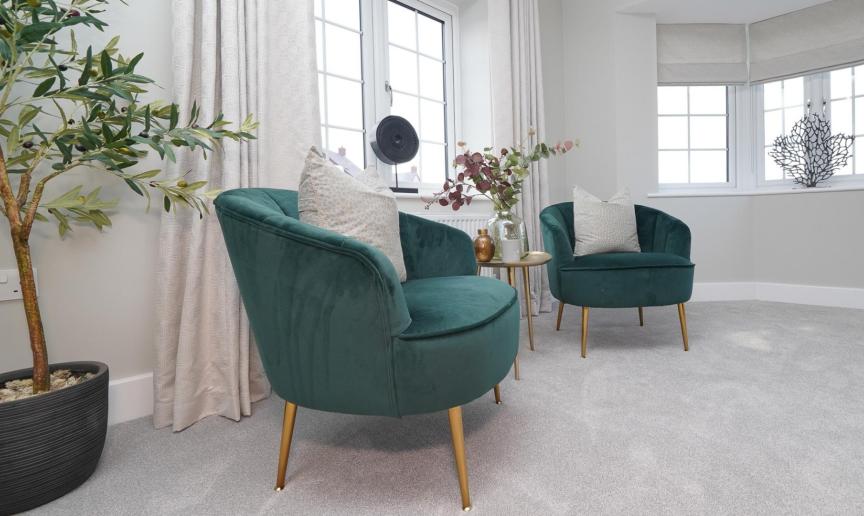 Two teal armchairs and a small side table in a bright, contemporary room with large windows, neutral décor, and a potted olive tree.