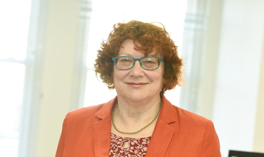 Professional portrait of a person wearing an orange blazer and patterned top, standing indoors in front of softly lit windows and facing the camera.