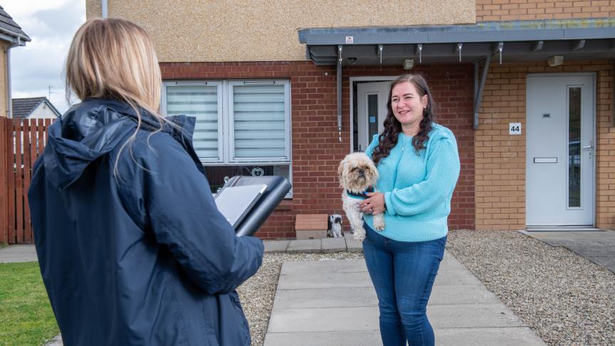 A person outside the front of their home holding a small dog facing another person who looks as though they are working for an organisation with a folder in their hand