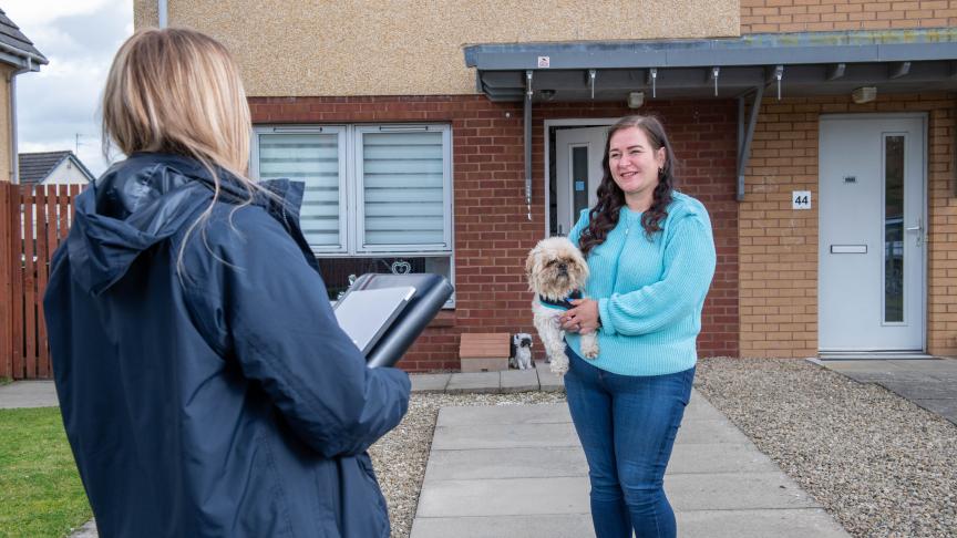 Who we say we are news image A person outside the front of their home holding a small dog facing another person who looks as though they are working for an organisation with a folder in their hand