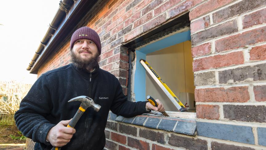 A man in a navy Sanctuary-branded fleece holds a chisel and hammer in front of a window where the double-glazing has been removed