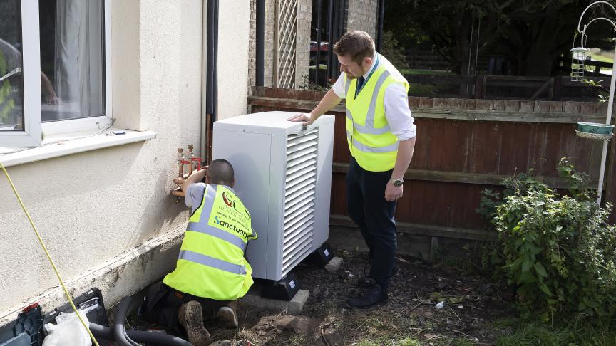 Two white, male Sanctuary staff members wearing high vis jackets are outside a building fitting a Swaffham heat pump