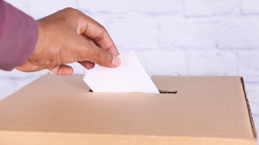 A hand holding a ballot paper posting it into the ballot box below
