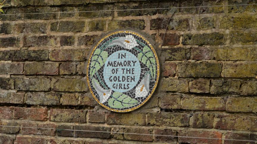 Circular mosaic plaque on a brick wall with the text 'IN MEMORY OF THE GOLDEN GIRLS', surrounded by white birds and green leaves