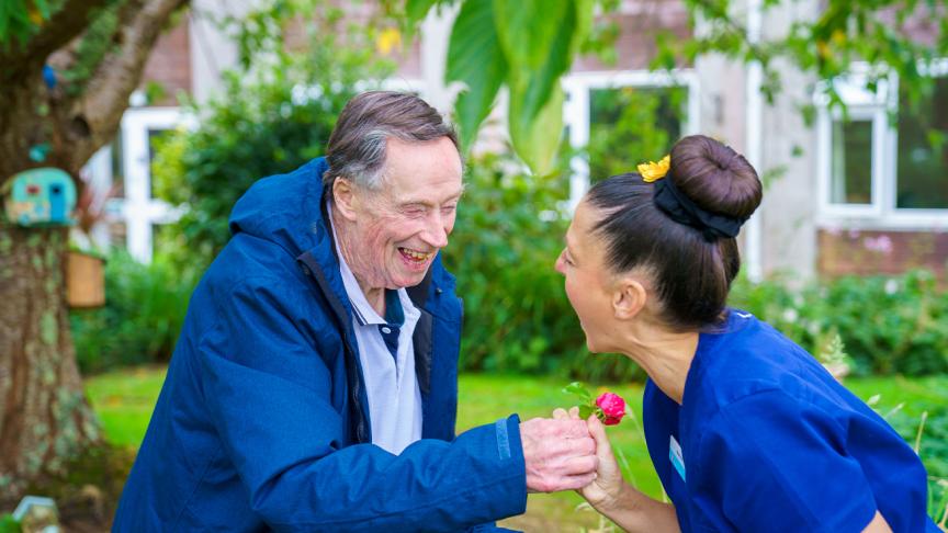 Person in blue jacket holds pink flower while another in blue scrubs leans forward; both are outdoors near a tree and building with greenery.