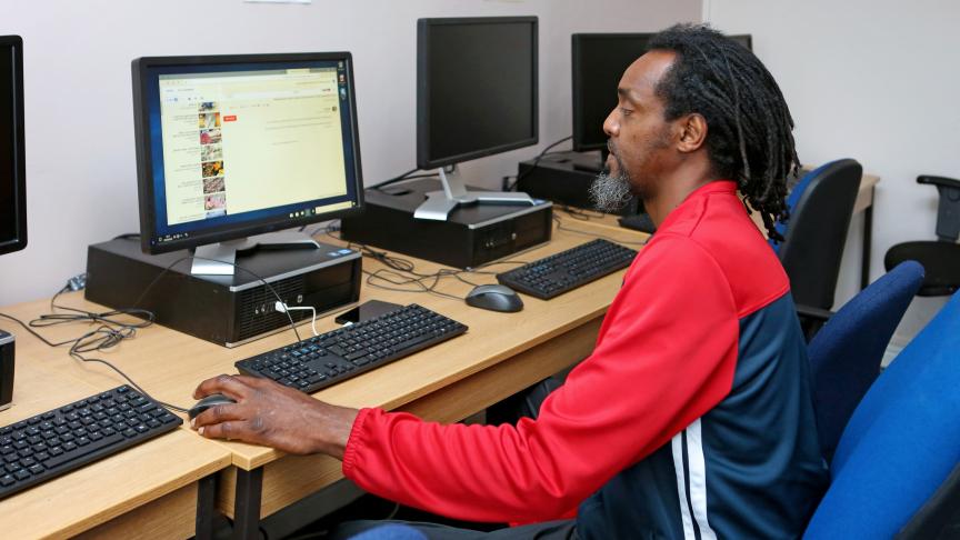 A man with long, curly hair and a beard is sitting at a computer desk in a room with multiple computers. He is wearing a red and black athletic jacket and is focused on the screen in front of him, which displays a web browser. The workspace is simple, with wooden desks and blue chairs.
