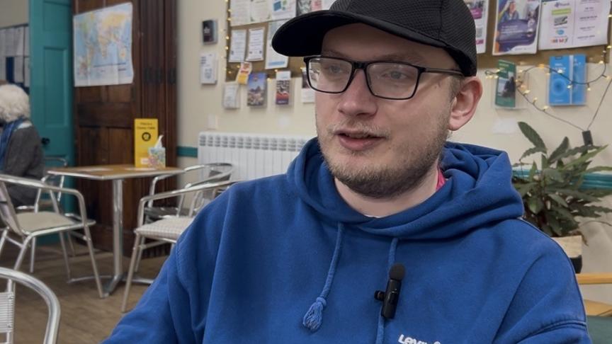 Person in blue Levi’s hoodie and black cap sitting in a café with bulletin board and chairs in background.