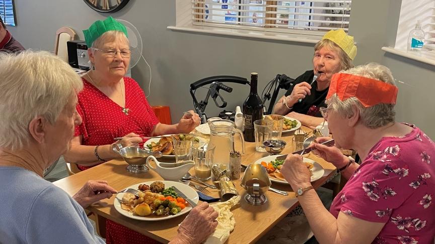 A group of four elderly women sitting around a table enjoying a meal together. They are wearing colorful paper crowns and are engaged in conversation.