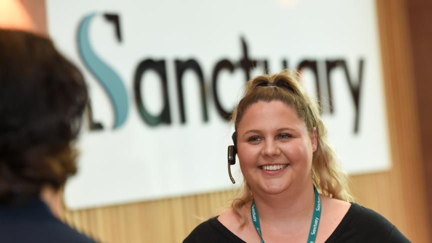 A smiling woman with curly blonde hair is wearing a headset and standing in front of a sign that reads "Sanctuary."