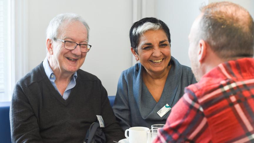 Three people sit around a small table, engaged in conversation with warm smiles. Cups and plates are on the table in a bright indoor setting.