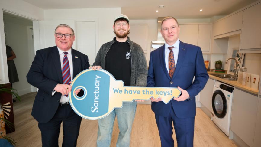 Three men are standing in a modern kitchen. The man in the middle is holding a large, symbolic key with the word "Sanctuary" and the phrase "We have the keys!" printed on it.