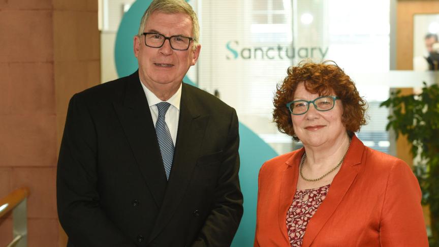 Two people standing side by side in an office reception area, with a large Sanctuary logo visible behind them.