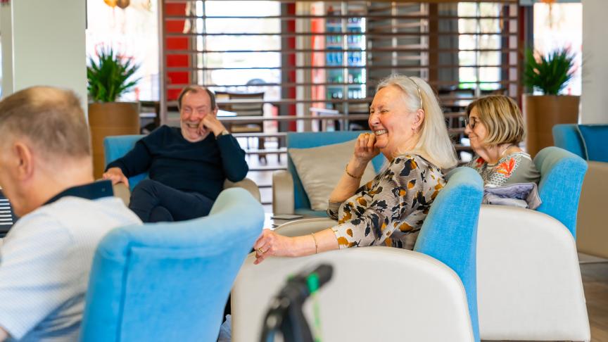 A group of three people is seated in a modern, brightly lit lounge area. In the foreground, a smiling older woman with long gray hair is laughing, wearing a patterned blouse. Beside her, a young girl with short hair is also smiling, while an older man in the background appears to be enjoying a humorous moment. The seating is comfortable, with light blue chairs, and the space is decorated with plants and warm lighting, creating a welcoming atmosphere.