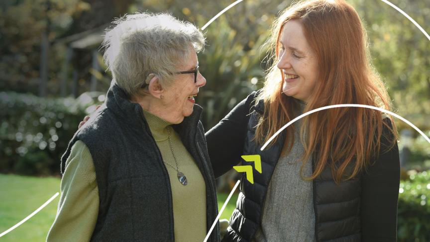 Two people standing together outdoors in a garden, with one person placing an arm around the other's shoulder.