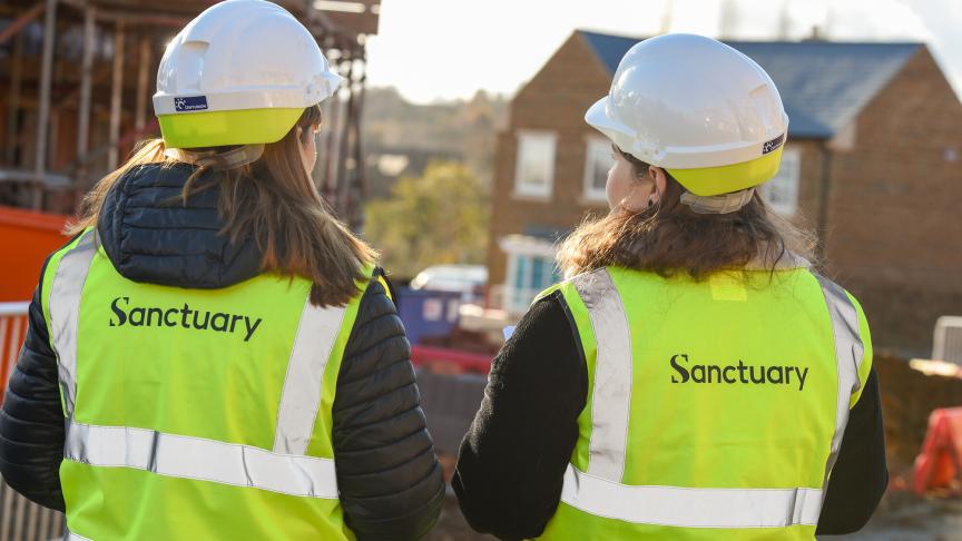 Two women wearing safety helmets and high-visibility vests with the word "Sanctuary" printed on the back are standing with their backs to the camera. They are on a construction site, with scaffolding and partially constructed buildings visible in the background. The scene is illuminated by sunlight, suggesting a clear day.