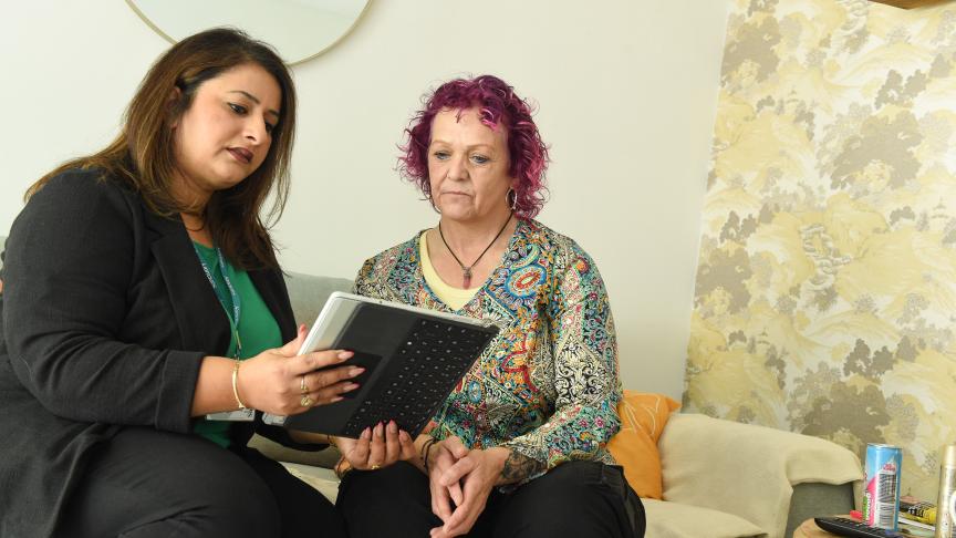 Colleague sits with a customer in a living room, using a tablet to review information together. The customer is seated on a sofa while the colleague points to items on the screen during a home visit.
